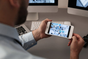 Wall Mural - Male security guard monitoring home cameras using smartphone indoors, closeup