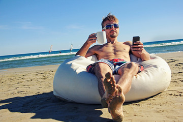 Attractive man in sunglasses and swimming pants sitting on pouf on beach with cup of tea or coffee and smartphone, in the background windsurfers in the sea. Active lifestyle