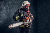 Fototapeta Do pokoju - Firefighter in safety helmet and oxygen mask holding a chainsaw. Studio photo against a dark textured wall