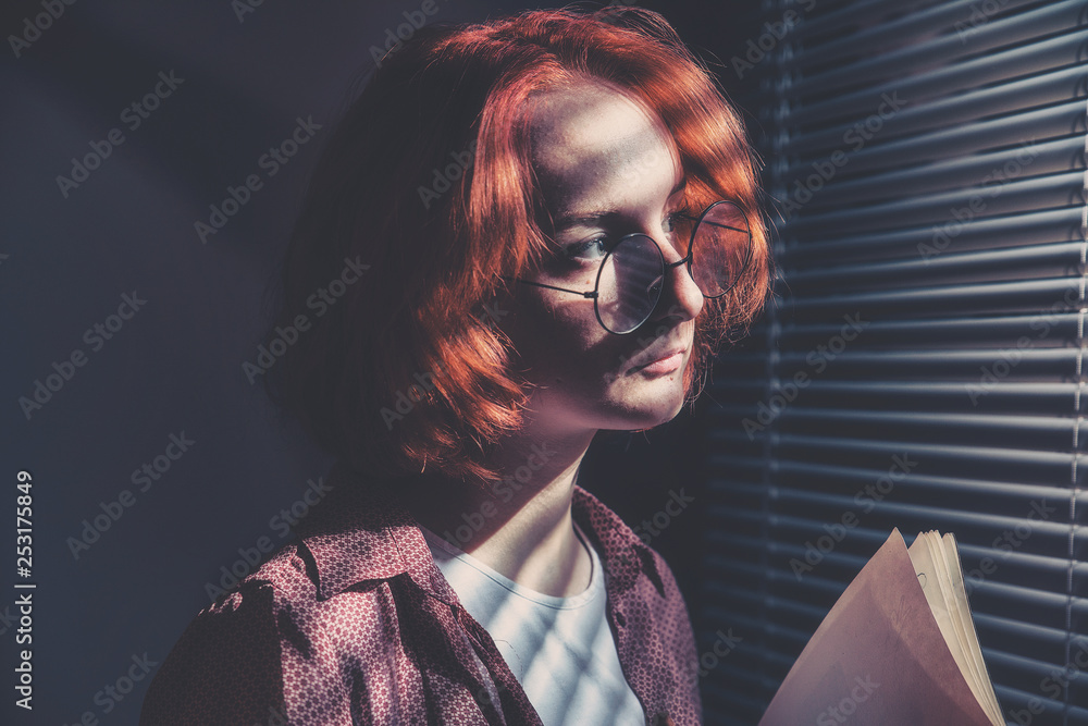 Red-haired girl with glasses in a plaid shirt and a white t-shirt is sad at the window with blinds - obrazy, fototapety, plakaty 