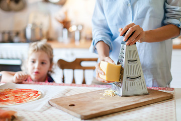 Wall Mural - Mother is cooking with child. Woman grates cheese with grinder. Family are preparing pizza and homemade italian food in kitchen. Lifestyle, authentic, candid moment.