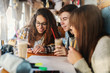 © dusanpetkovic1 - Three smiling multicultural classmates sitting at coffee shop, drinking coffee and looking at smart phone. On desk notebooks, markers and pens.