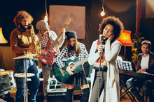 Mixed Race Woman Singing. In Background Band Playing Instruments. Home Studio Interior.