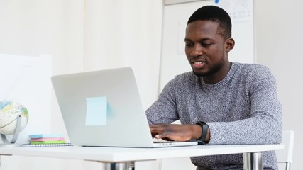 Medium shot smiling young African programmer using laptop at workplace in modern office. Low angle busy man writing program code for startup application on notebook pc