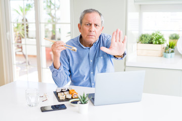 Wall Mural - Handsome business senior man eating delivery sushi while working using laptop with open hand doing stop sign with serious and confident expression, defense gesture