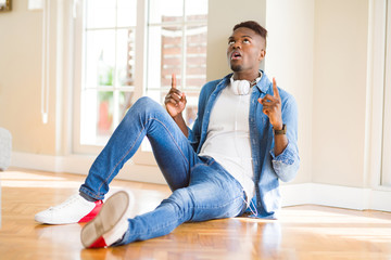 Poster - African american man wearing headphones listening to music sitting on the floor amazed and surprised looking up and pointing with fingers and raised arms.