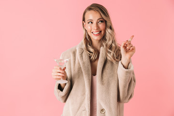 Emotional young pretty woman posing isolated over pink wall background holding glass with alcohol drink.