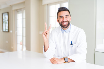 Handsome hispanic doctor or therapist man wearing medical coat at the clinic showing and pointing up with fingers number two while smiling confident and happy.