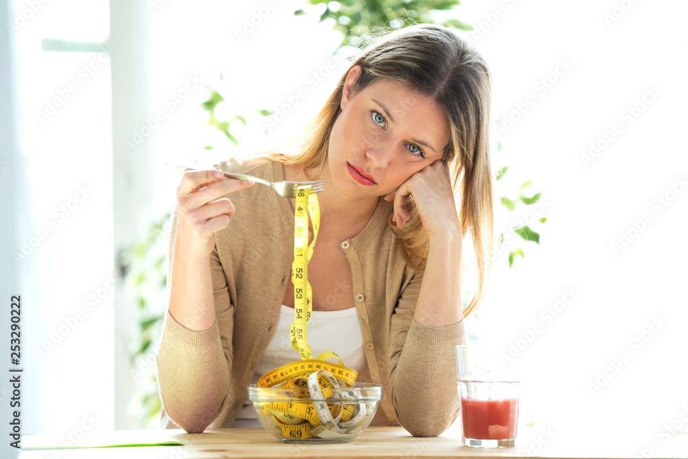 Worried beautiful woman holding fork with measuring tapes sitting in living room at home. - obrazy, fototapety, plakaty 
