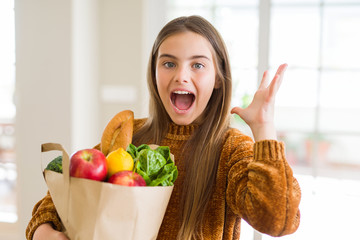 Wall Mural - Beautiful young girl holding paper bag of fresh groceries very happy and excited, winner expression celebrating victory screaming with big smile and raised hands