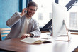 © opolja - Young smiling bearded businessman is sitting at table in front of computer, talking on cell phone. Freelancer, entrepreneur works at coworking centre. Telephone conversations.