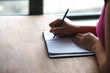 © Joseph Kirsch - A closeup photograph of mixed race woman wearing a pink shirt and holding a blue pen writing in a modern style note pad sitting at a wood dining table or desk with morning light coming from windows.