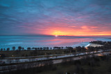 Fototapeta Niebo - A beautiful sunrise landscape photograph of the bright orange and yellow sun peering over the horizon on Lake Michigan in Chicago with pink and blue clouds in the sky and cars on Lake Shore Drive.