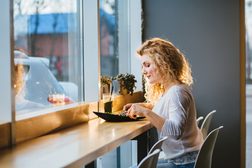 Wall Mural - Curly blond girl have a dinner. Charming woman eating salad alone during rest in coffee shop, happy Caucasian female have a dinner while relaxing in cafe during free time.