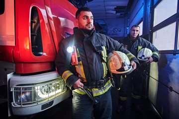 Wall Mural - Two firemen wearing protective uniform standing next to a fire engine in a garage of a fire department.