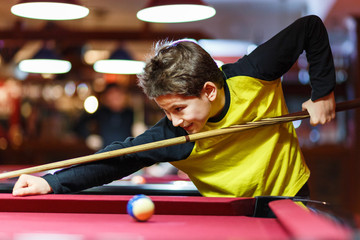 Cute boy in yellow t shirt plays billiard or pool in club. Young Kid learns to play snooker. Boy with billiard cue strikes the ball on table. Active Leisure, sport, hobby concept