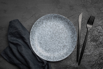 Stylish table setting with black cutlery. Empty plate, table textile and black cutlery on black concrete background. Top view. Restaurant, menu, cafe or food concept