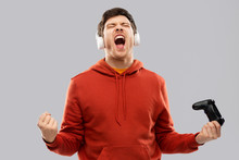 Technology, Gaming And People Concept - Happy Young Man Or Gamer In Headphones With Gamepad Winning In Video Game