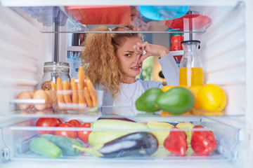 Wall Mural - Woman standing in front of opened fridge and holding up to her nose because of bad smell. Picture taken from the inside of fridge full of groceries.