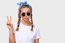 Studio Portrait Of Cheerful Blonde Young Woman Wearing Trendy Sunglasses, White T-shirt And Blue Headband, Making A Duck Face And Showing Peace Sign. Student Girl Going Crazy With Braids Hairstyle