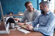 © opolja - Shot of two business colleagues working in their office using a desktop computer. Man sitting at his desk with male colleague sitting by