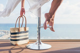 Fototapeta Natura - A girl with a beach bag is sitting on a bar stool in a summer cafe on the sea and sky background. Bottom view on barefoot female legs