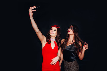 Wall Mural - Photo of two young party women smiling taking selfie photo on mobile phone over black background