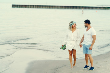 elegant and stylish woman with light curly hair dressed in a white blouse walking on the beach near water along with her elegant man in a white t-shirt and in a  blue shorts