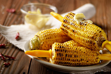 Wooden table with deep grilled sweet corn cobs under melting butter with plastic holder on clay dish