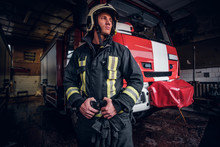 Portrait Of A Brave Young Fireman Wearing Protective Uniform Standing Next To A Fire Engine In A Garage Of A Fire Department