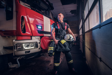 Wall Mural - Muscular fireman holding a protective helmet in a garage of a fire department, standing next to a fire engine and looking sideways