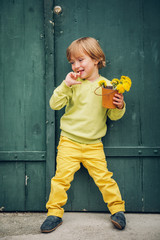 Wall Mural - Outdoor portrait of a cute little boy, wearing yellow pullover and trousers and blue moccasins, standing by the green wooden door, holding bucket with flowers dandelions