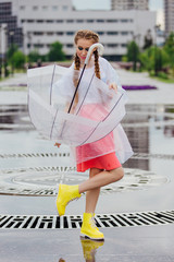 Young pretty girl with two braids in yellow boots and with transparent umbrella stands near fountain.