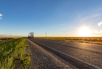 Wall Mural - A truck travels along an empty long straight road in the New South Wales countryside on a blue sky day at sunset.