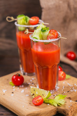 Two glasses of tomato juice decorated with fresh tomatoes, cucumber and leaves on a wooden background