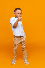 Wall Mural - Full length image of confident cool dark skinned schoolboy in fashionable wear standing at yellow studio wall, pointing fore finger at camera, winking at you. Beauty, elegance, style and fashion