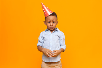 Wall Mural - Picture of frustrated unhappy Afro American little boy wearing red cone hat having mournful upset facial expression, making nervous gesture after being told by his father for misbehavior at party