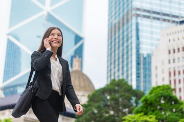 Wall Mural - Asian businesswoman talking on mobile phone walking in Hong Kong city street to office, skyscrapers cityscape background. Young woman on smartphone happy. Multiracial Chinese Caucasian lady.