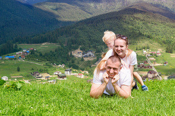 Happy family: joyful father, mother and two sons are lying on the green grass against the background of the forest, mountains and sky with clouds.