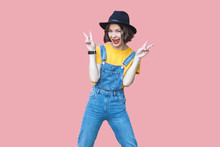 Portrait Of Excited Beautiful Young Woman In Yellow T-shirt And Blue Denim Overalls With Makeup And Black Hat Standing And Looking With Victory Or Peace Sign. Studio Shot Isolated On Pink Background.
