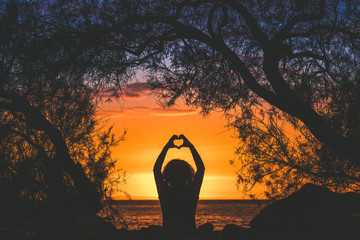 Silhouette image of a young woman making heart with hands on a beautiful summer evening Back view of a girl fingers like hearts sunset in background and branches that frame Enjoyable romance feelings