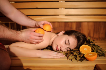 Wall Mural - lateral view of a beautiful woman enjoying massage procedure with oranges and birch broom in a wooden sauna