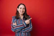 Smiling Woman In  Casual Clothes With Arm On Hip Pointing Away While Looking At The Camera Over Red Background