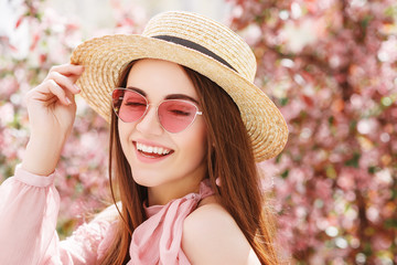 Outdoor close up portrait of young beautiful happy smiling woman wearing stylish pink cat eye sunglasses, straw hat, earrings, blouse, posing in street of city. Copy, empty space for text