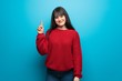 © luismolinero - Woman with red sweater over blue wall showing and lifting a finger in sign of the best