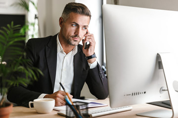 Wall Mural - image of serious businessman talking on cell phone while working on computer in office
