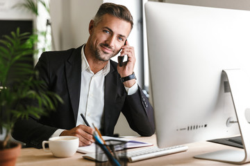 Image of happy businessman talking on cell phone while working on computer in office