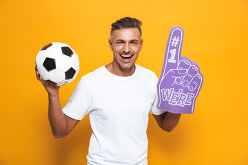 Wall Mural - Image of beautiful man 30s in white t-shirt holding soccer ball and number one fan hand glove with finger raised while standing isolated