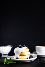 Cottage Cheese Pancakes One On Another Topped With Cream, Blueberries And Sugar Powder And Cup Of Coffee, Fork And Green Mint Leaves On Black Background, Vertical With Copy Space For Text Or Design