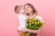 Young mother hugs his little son sitting on the floor against a pink background. Mom holds a bouquet of spring yellow flowers. Care and relationships and family concept. Mothers day